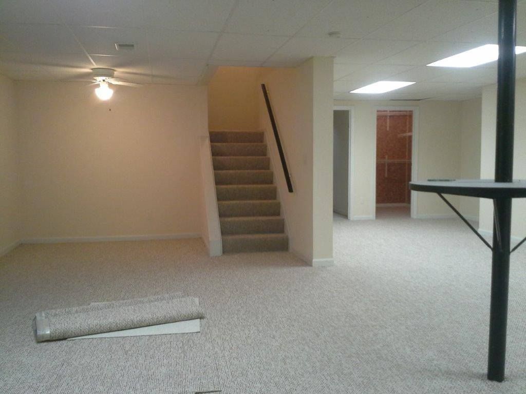 A spacious, carpeted basement room with stairs leading upward and a vertical support pole on the right.
