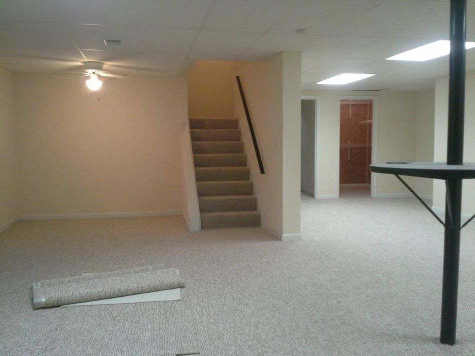 A carpeted basement with a staircase leading up, neutral walls, recessed ceiling lights, and a support pole on the right.
