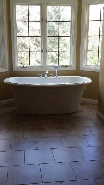 A white freestanding bathtub centered in front of a bay window in a room with brown tiled floors and warm-toned walls.