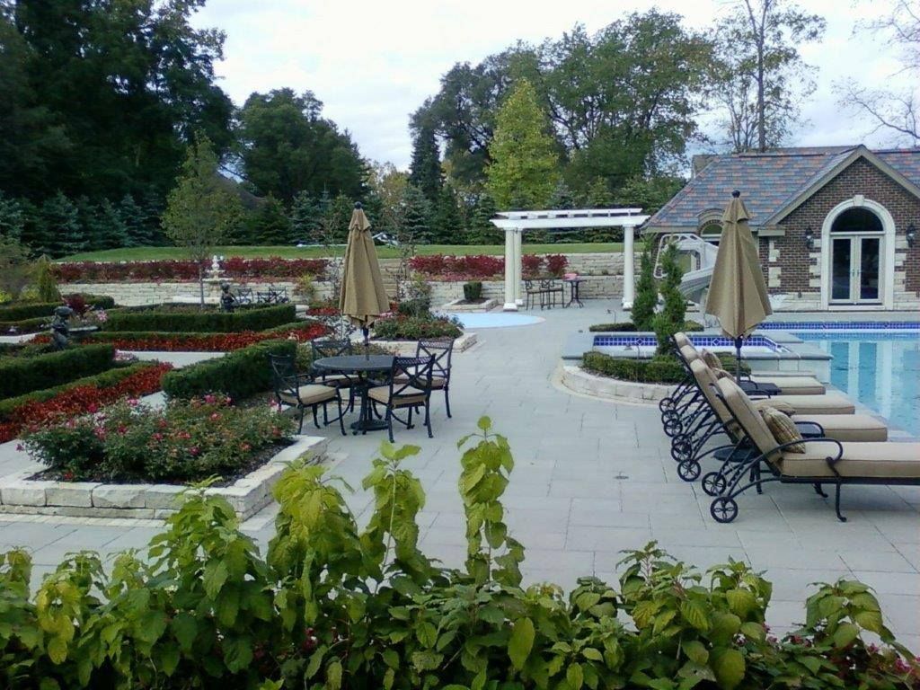 A backyard patio features a swimming pool, lounge chairs, a dining set under an umbrella, and manicured garden beds.