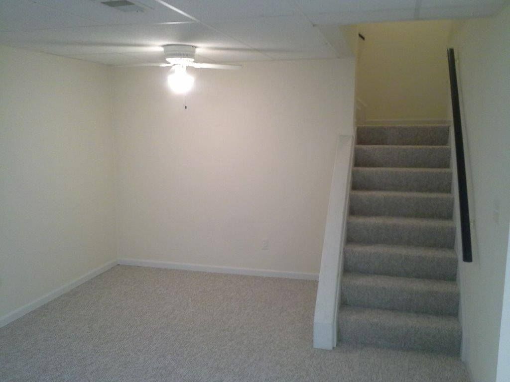 A carpeted room with light-colored walls and a ceiling fan, featuring a staircase leading upward to the right.