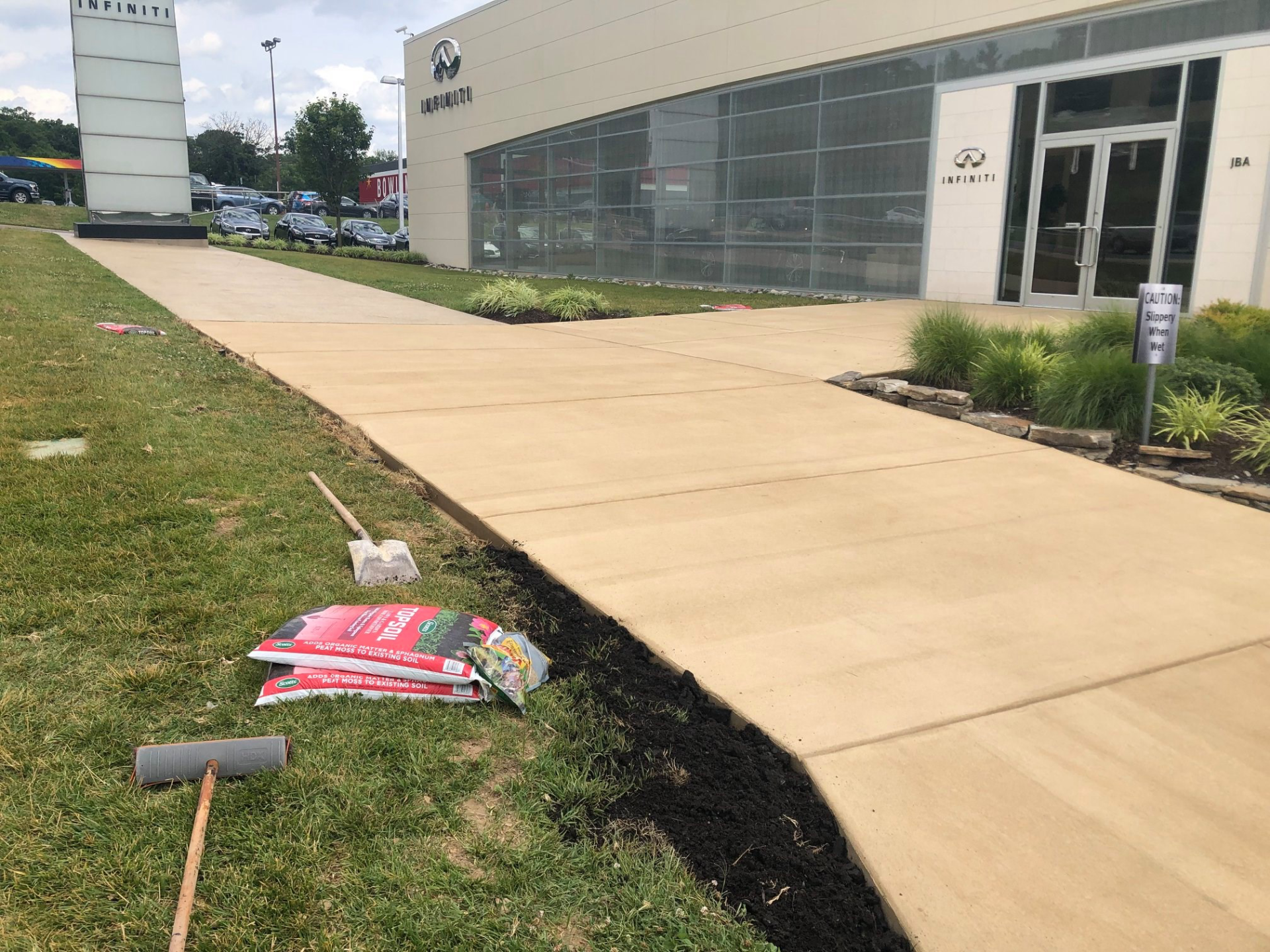 Sidewalk next to a building, with bags of mulch, a shovel, and a rake on the grass.
