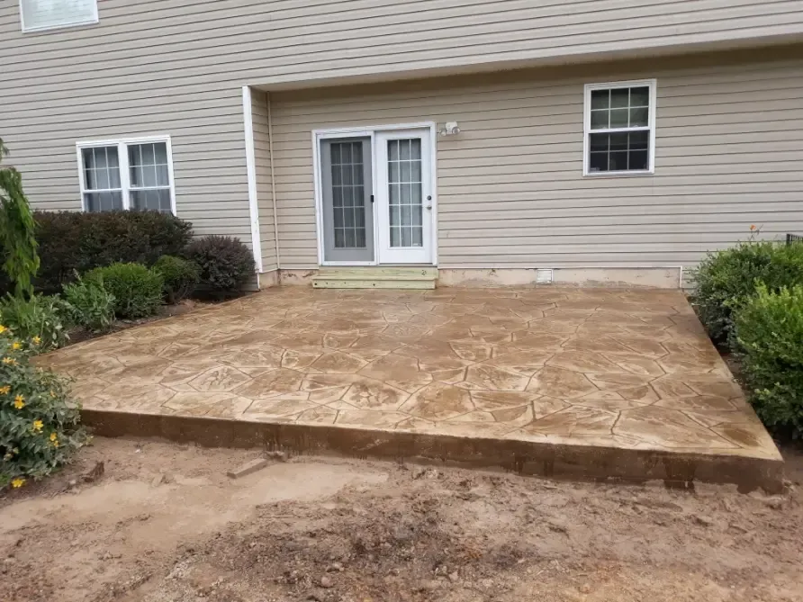 Backyard patio, textured brown concrete, next to house with sliding door and windows; surrounded by dirt and bushes.