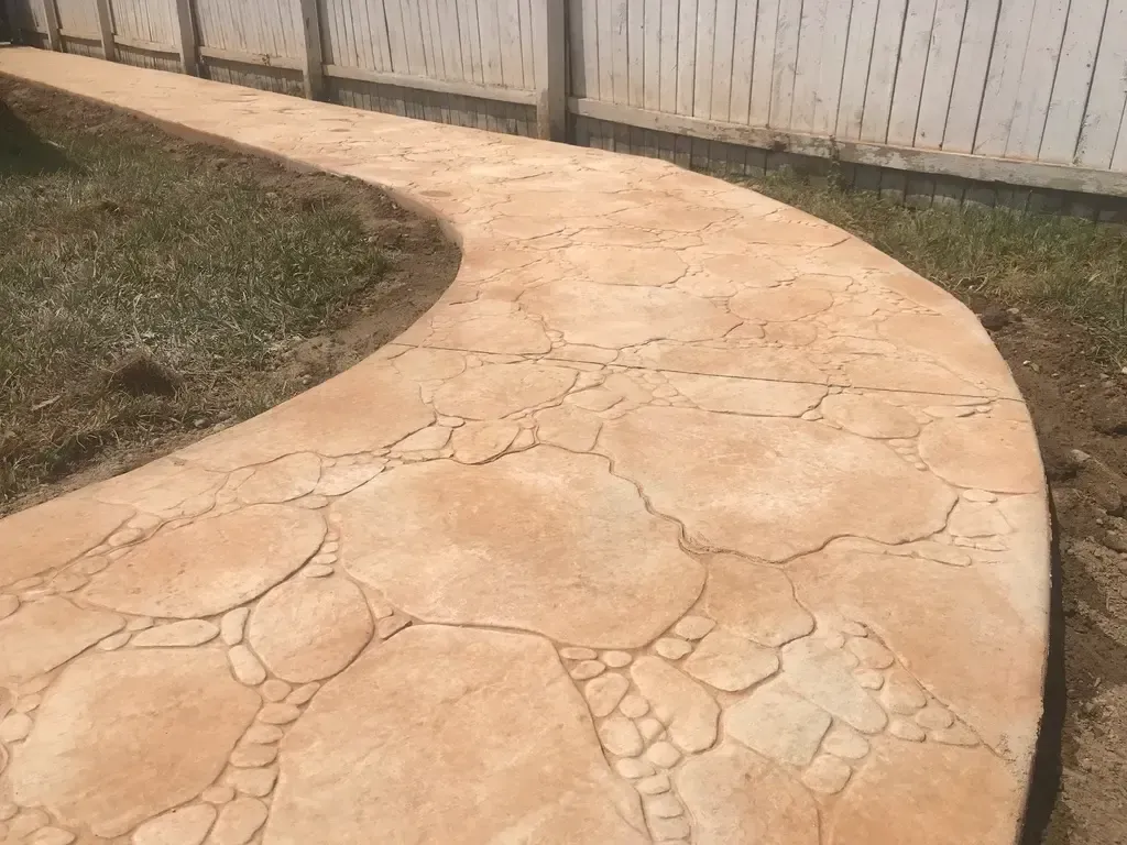 Curving stamped concrete walkway in a yard with a white fence and grass.