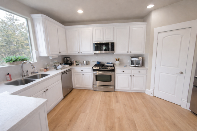 Bright white kitchen with wood floors, stainless appliances, and a large window over the sink.