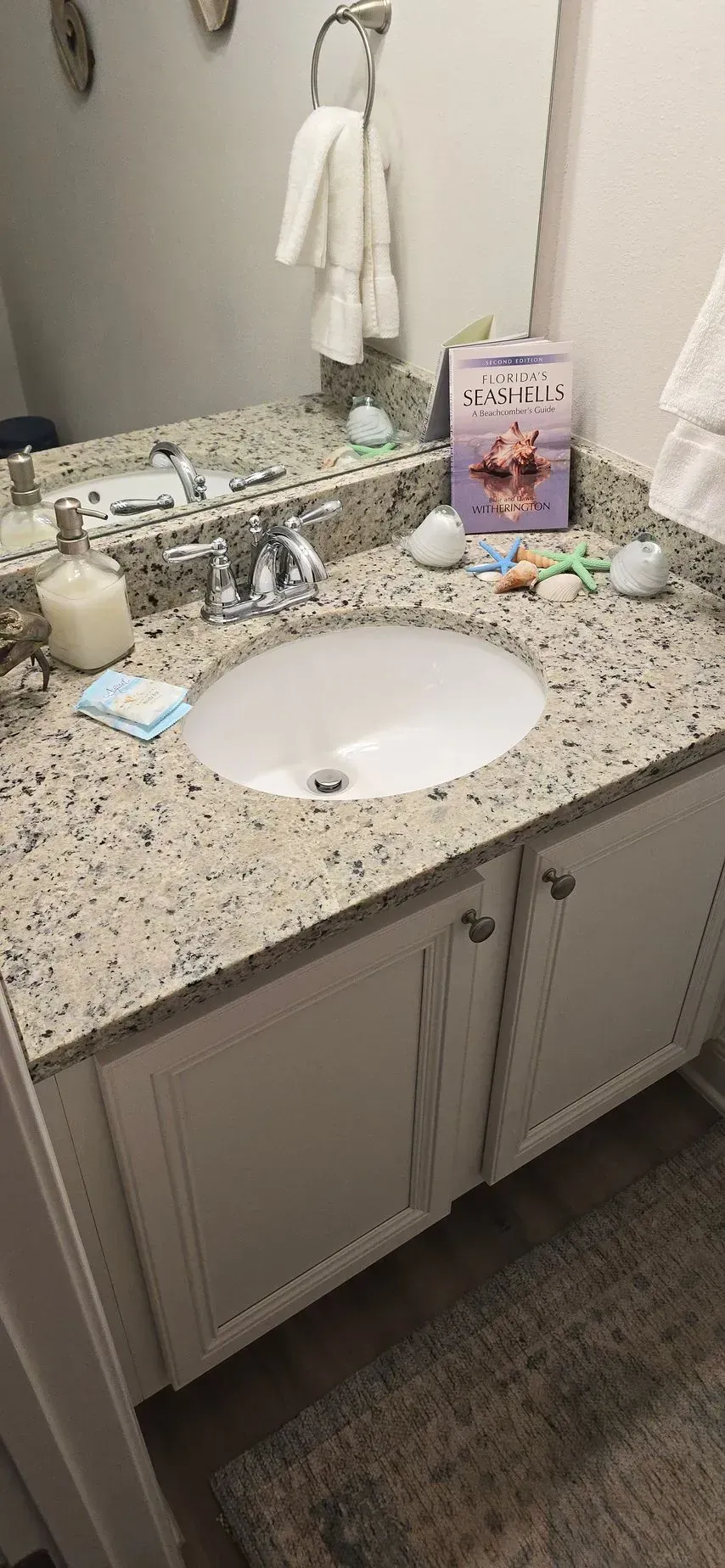 Modern, beige bathroom with a glass shower, toilet, and sink. Features include artwork and accessible grab bar.