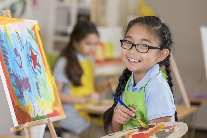 Girl with glasses smiles, holding a paintbrush and palette, painting on a canvas in an art class.