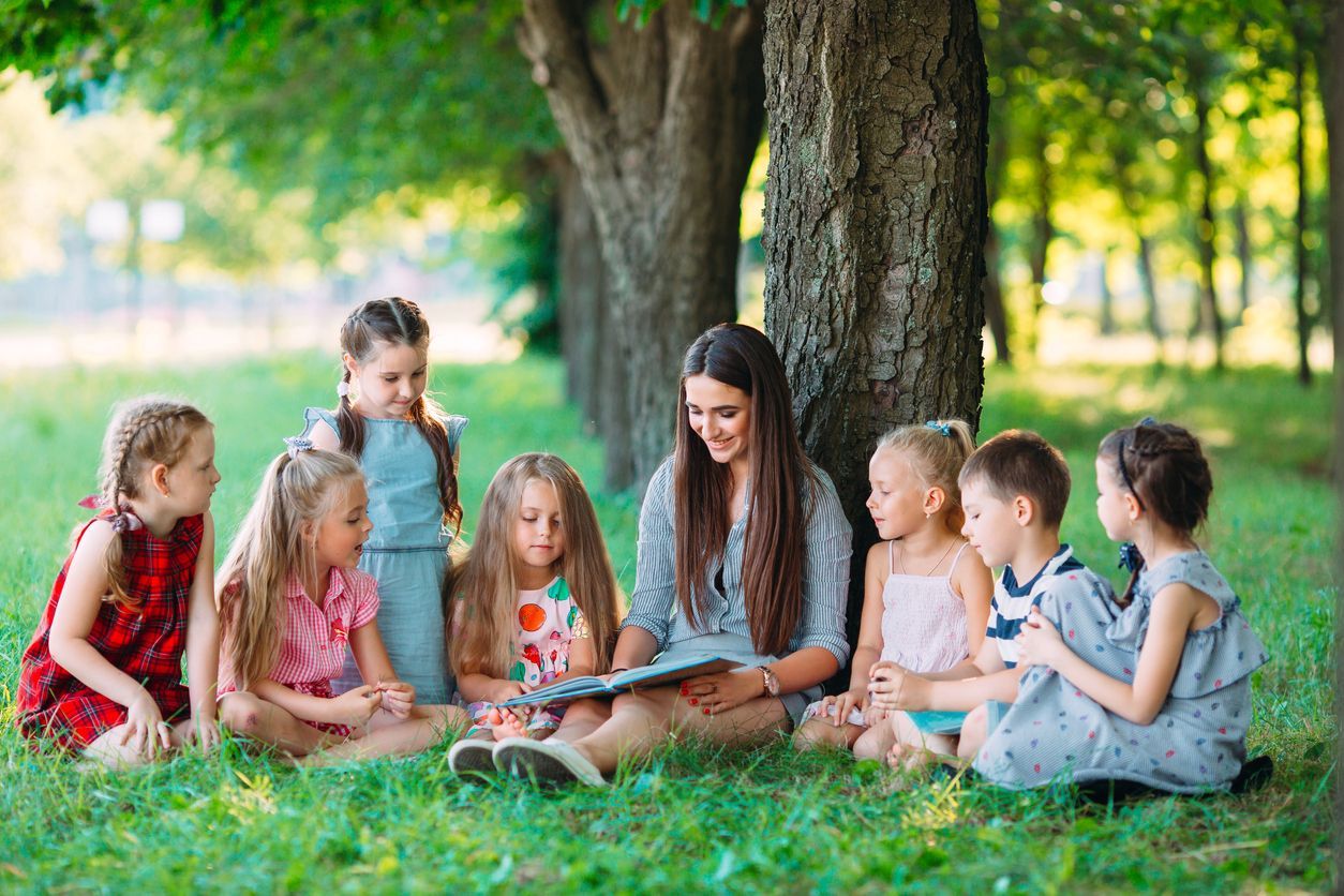 A woman reads a book to a circle of children sitting on grass under a tree.
