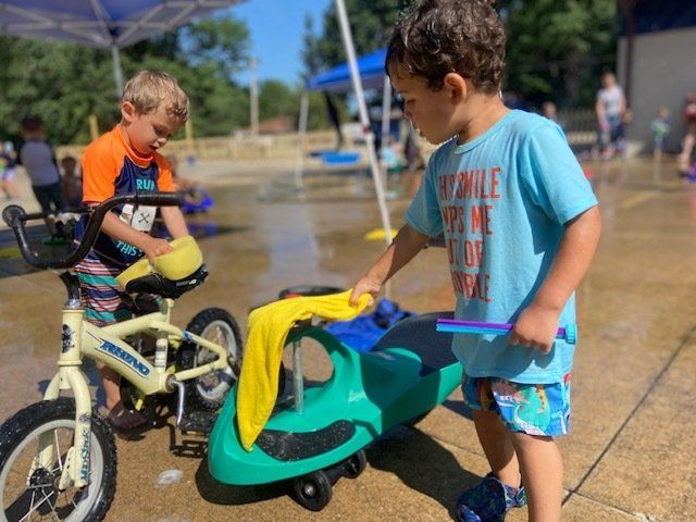 Two children washing a bicycle and a toy car in a splash pad.