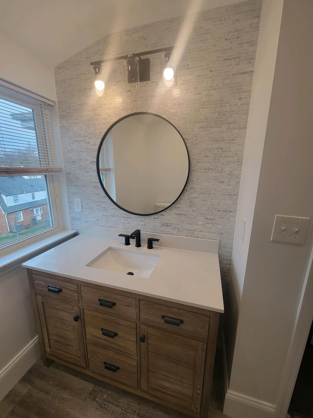 Bathroom with wooden vanity, round mirror, and textured gray wall tiles.