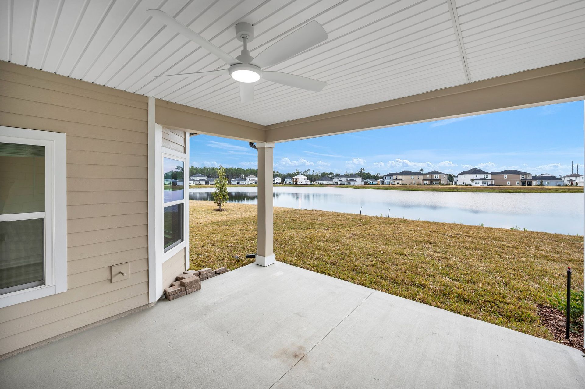 Covered patio overlooking a grassy yard and calm lake, with beige siding, white columns, and a ceiling fan.