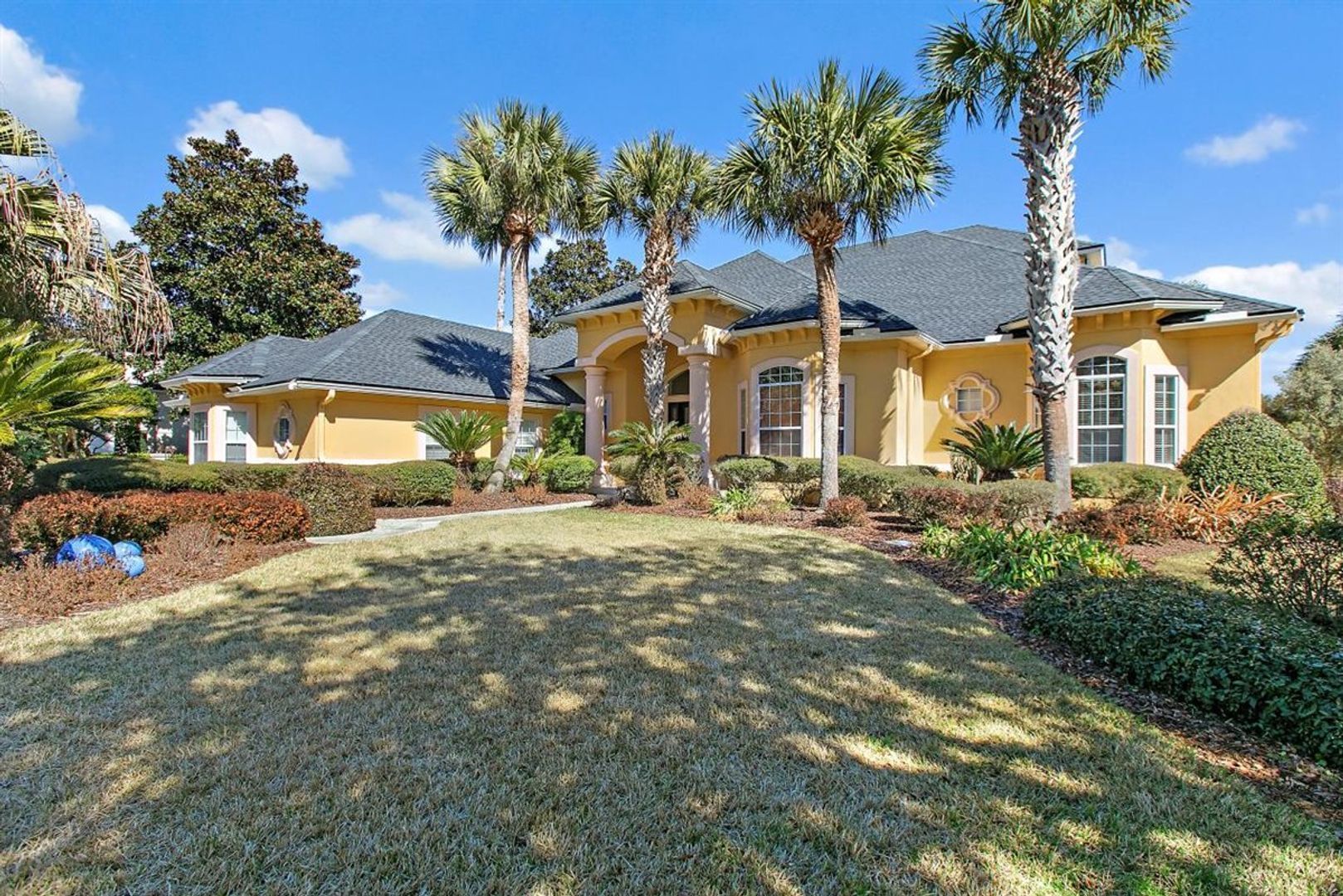 Large tan suburban house with a blue roof, palm trees, and a sloped front lawn under a clear sky