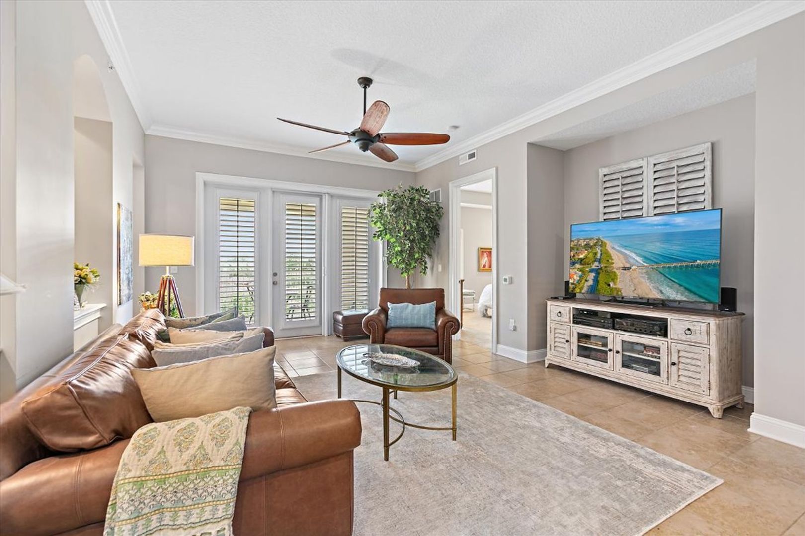 Bright living room with leather sofa, glass coffee table, TV console, ceiling fan, and plantation shutters.