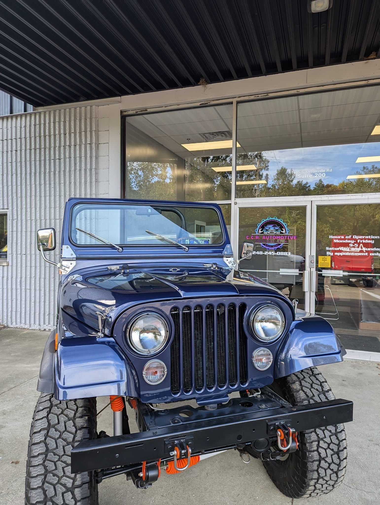 Blue vintage Jeep parked in front of a building with a black grill, black bumper, and orange accents.