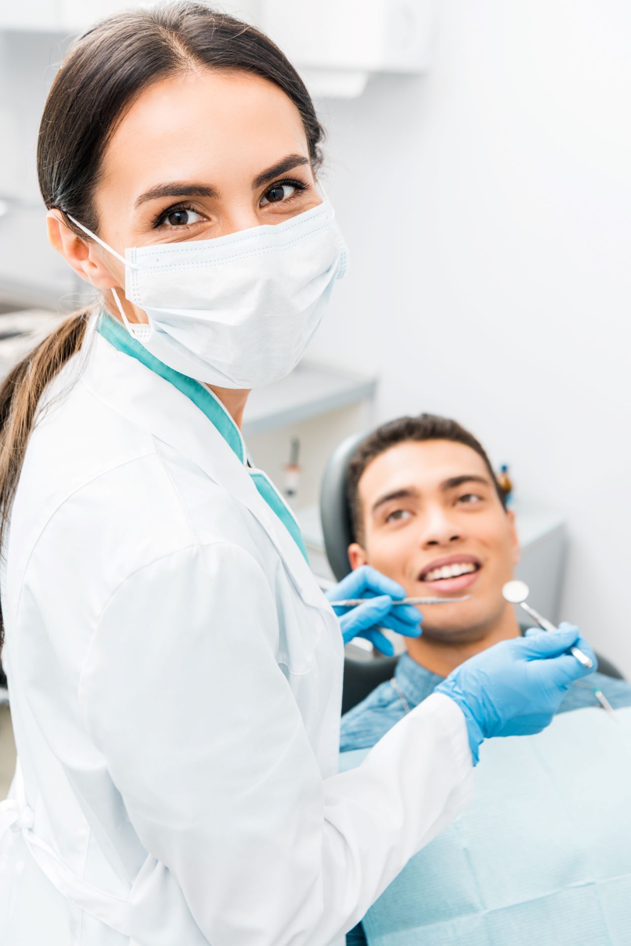 Dental clinician in white coat and mask treating a smiling patient in a dental chair