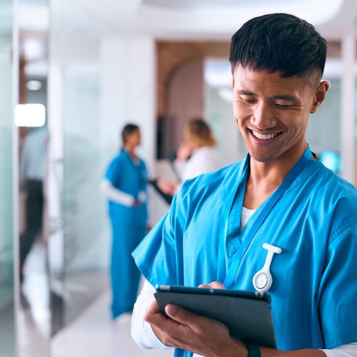 Smiling nurse in blue scrubs using a tablet in a hospital hallway