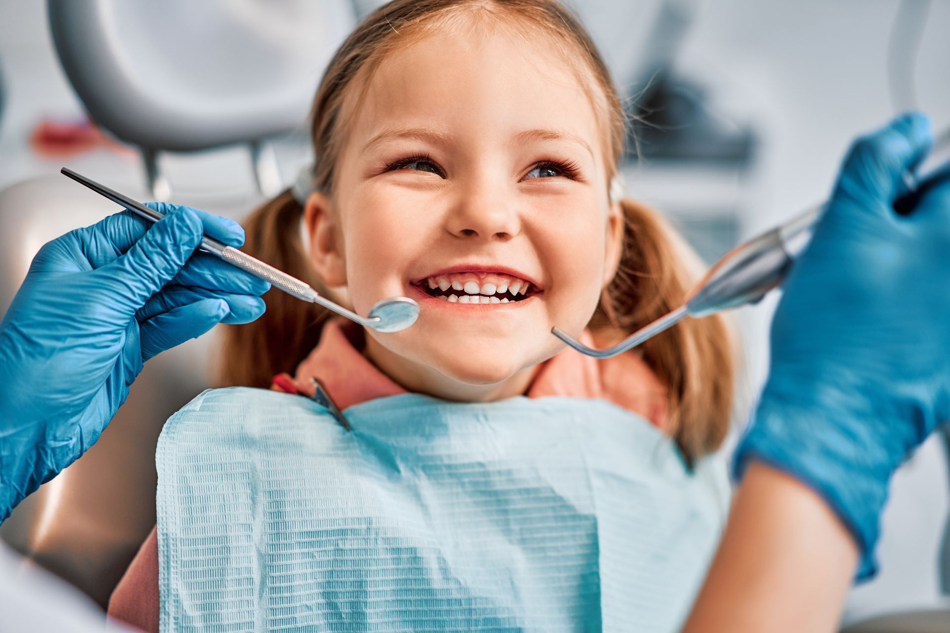 Child at the dentist with open mouth while gloved hands hold dental tools near a bib.