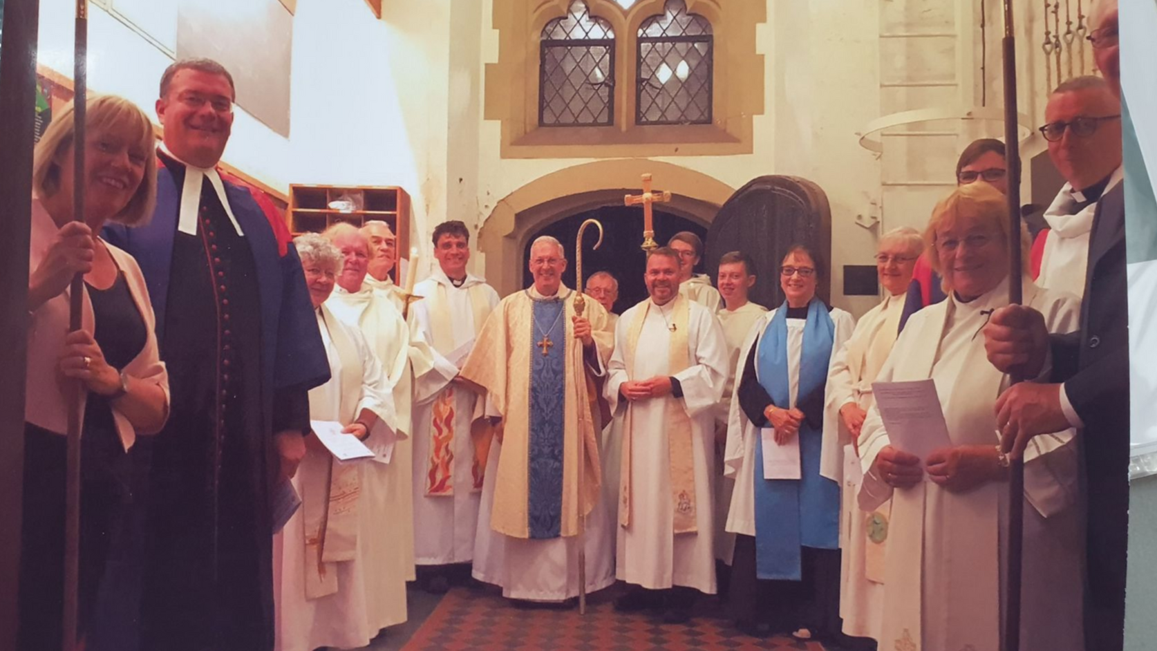 Clergy and parishioners in a church doorway; some hold banners and papers, all wearing white robes.