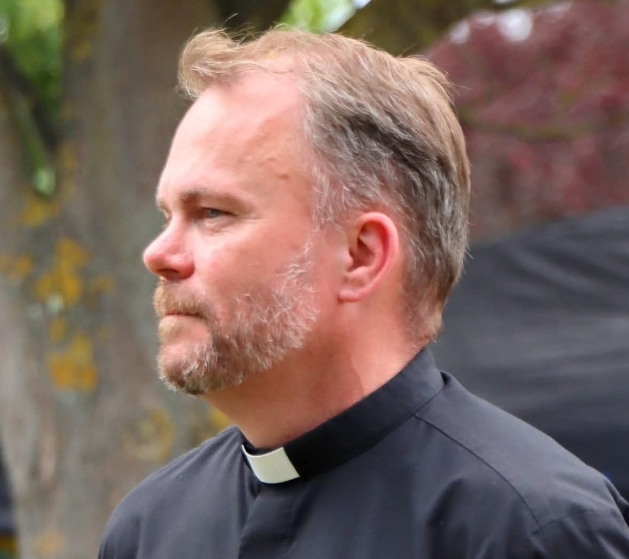 Man in black clerical shirt with white collar, looking left, outdoor setting.