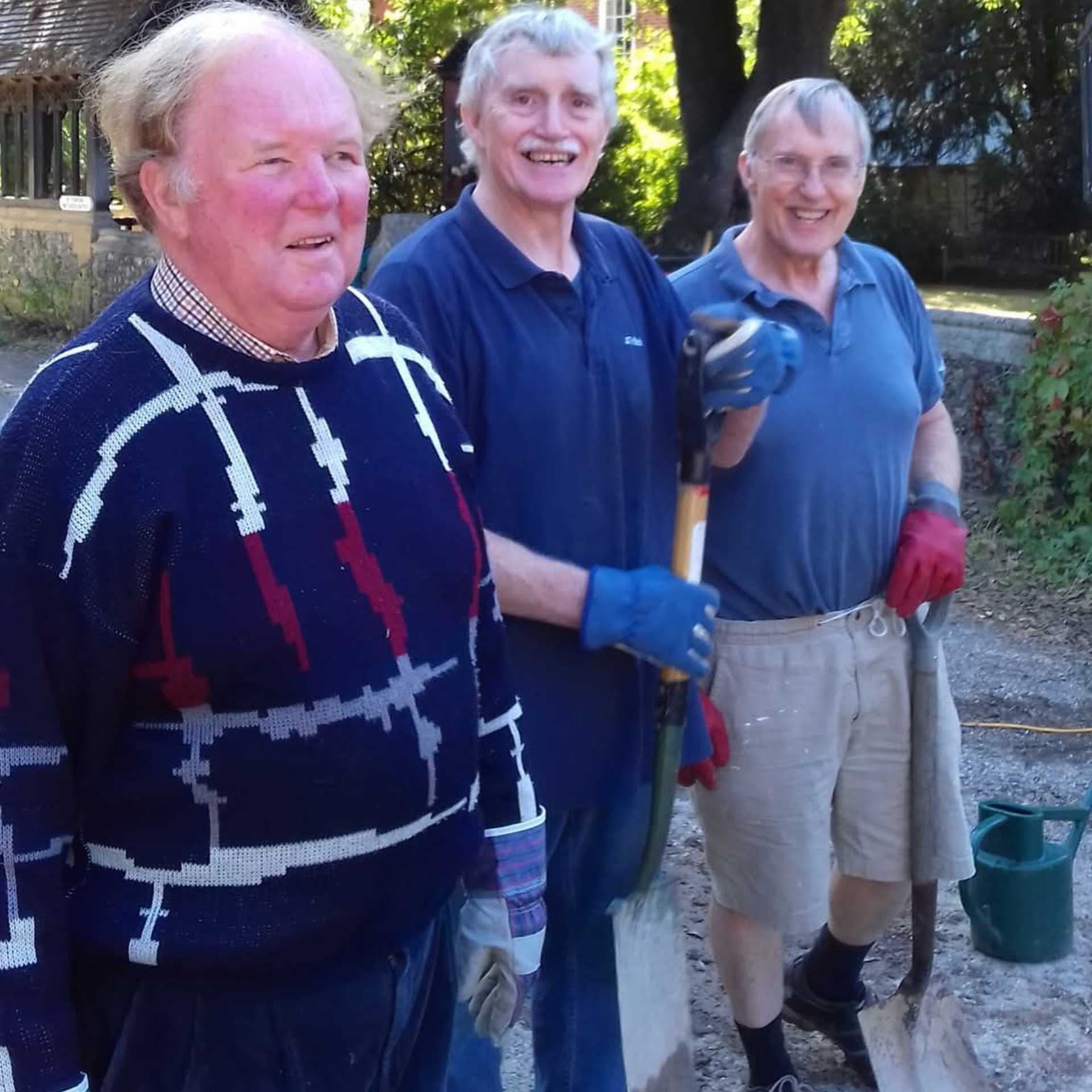 Three men holding shovels, standing outside; one wearing a patterned sweater, two in blue shirts.