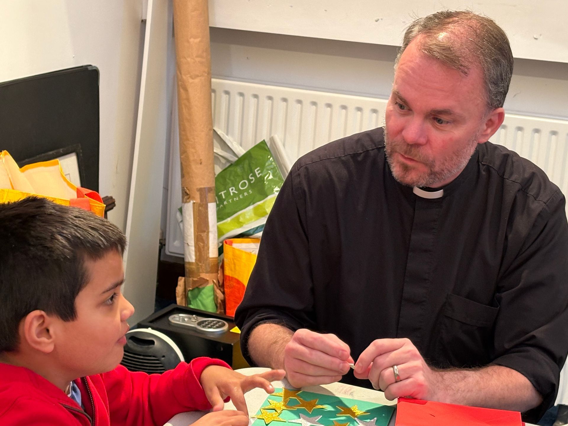 Clergyman seated with a child, both looking at a craft project on a table.