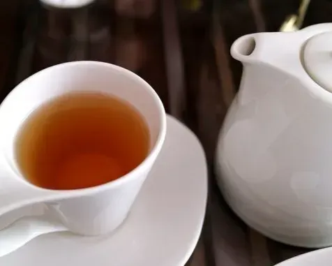White teacup with tea and saucer next to a white teapot on a striped table.