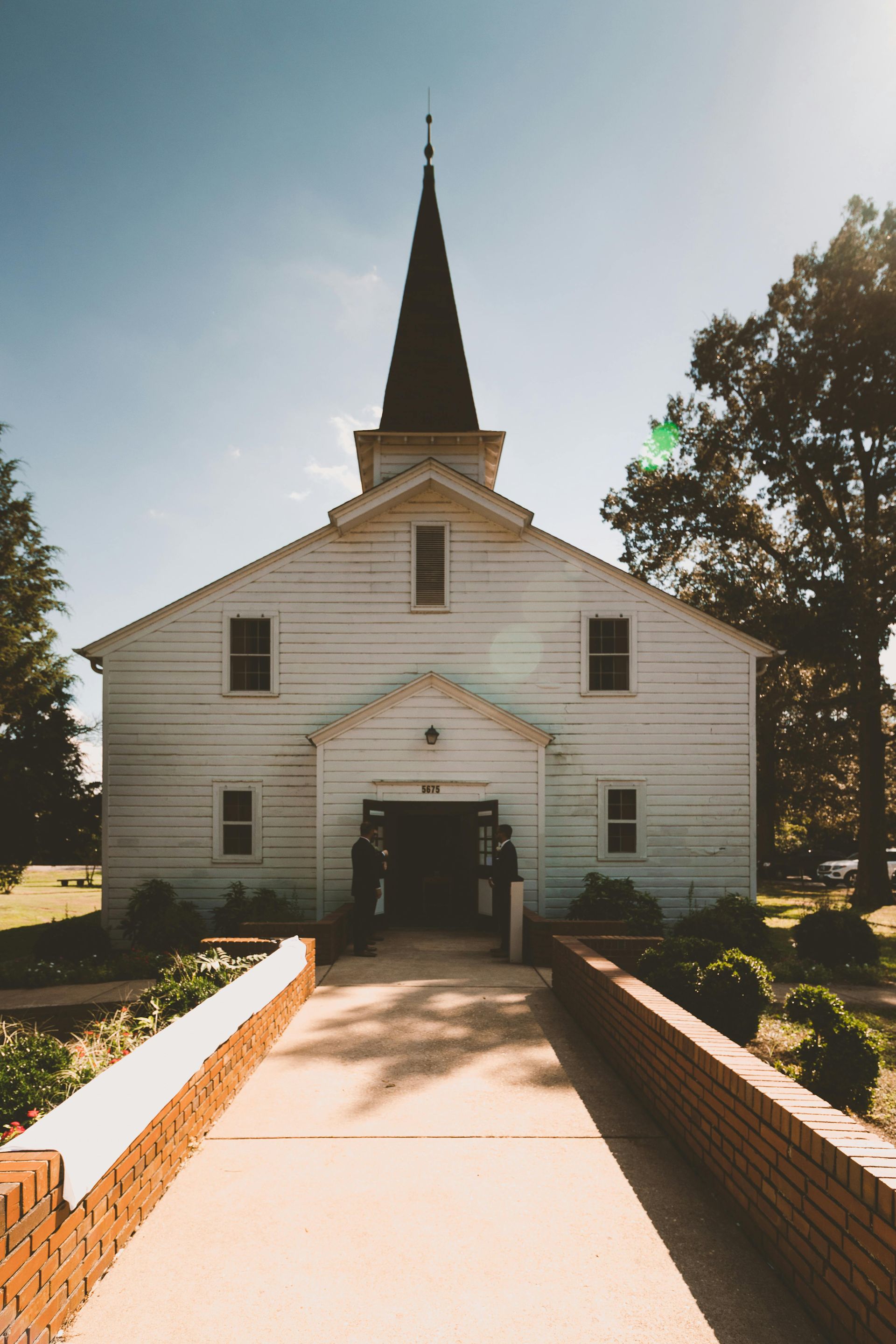 White church building with tall steeple, brick walkway leading to the entrance, sunny day.