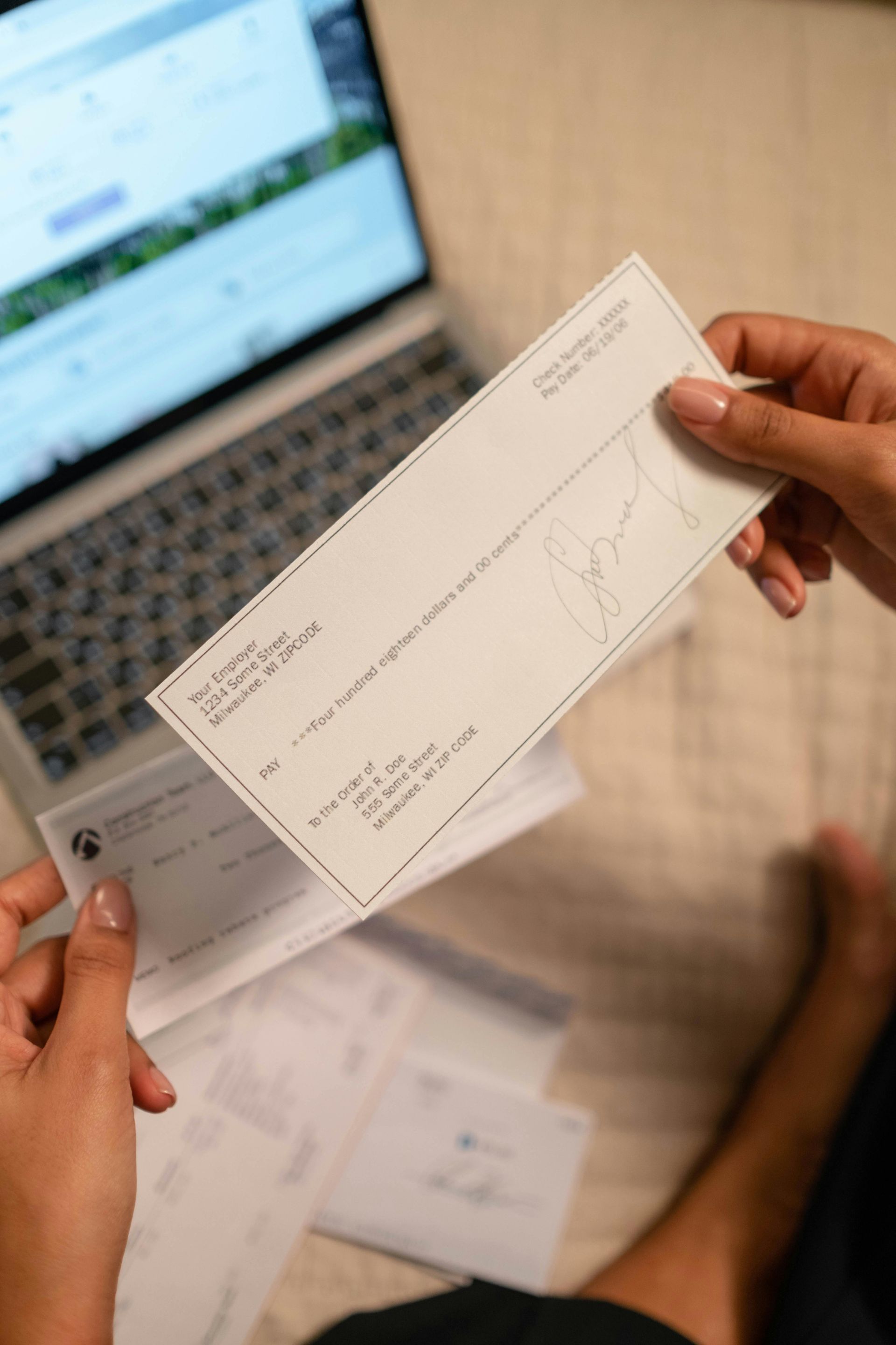 Hands holding a business check, laptop in the background. Other checks scattered below.