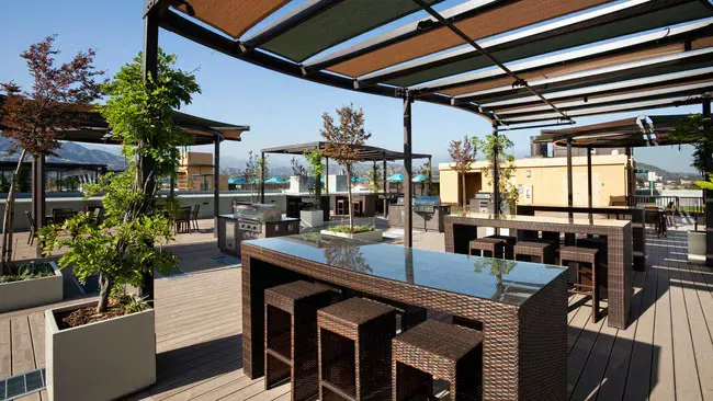 Rooftop patio with tables, chairs, shade structure, plants, and cityscape backdrop.