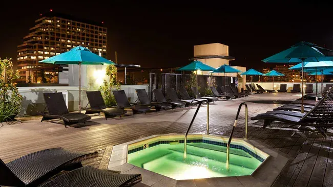 Rooftop pool area at night with lit hot tub, lounge chairs, and turquoise umbrellas. City skyline in the background.