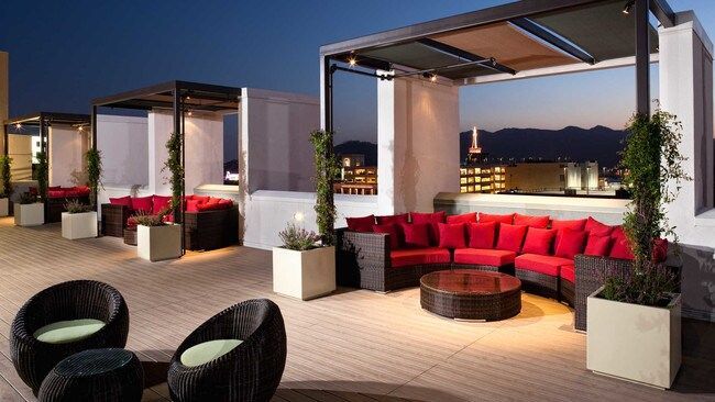 Rooftop lounge with red couches and potted plants; city skyline in the distance at dusk.