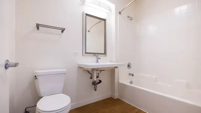 A small, white bathroom with a toilet, sink, mirror, and bathtub; features wood-like flooring.