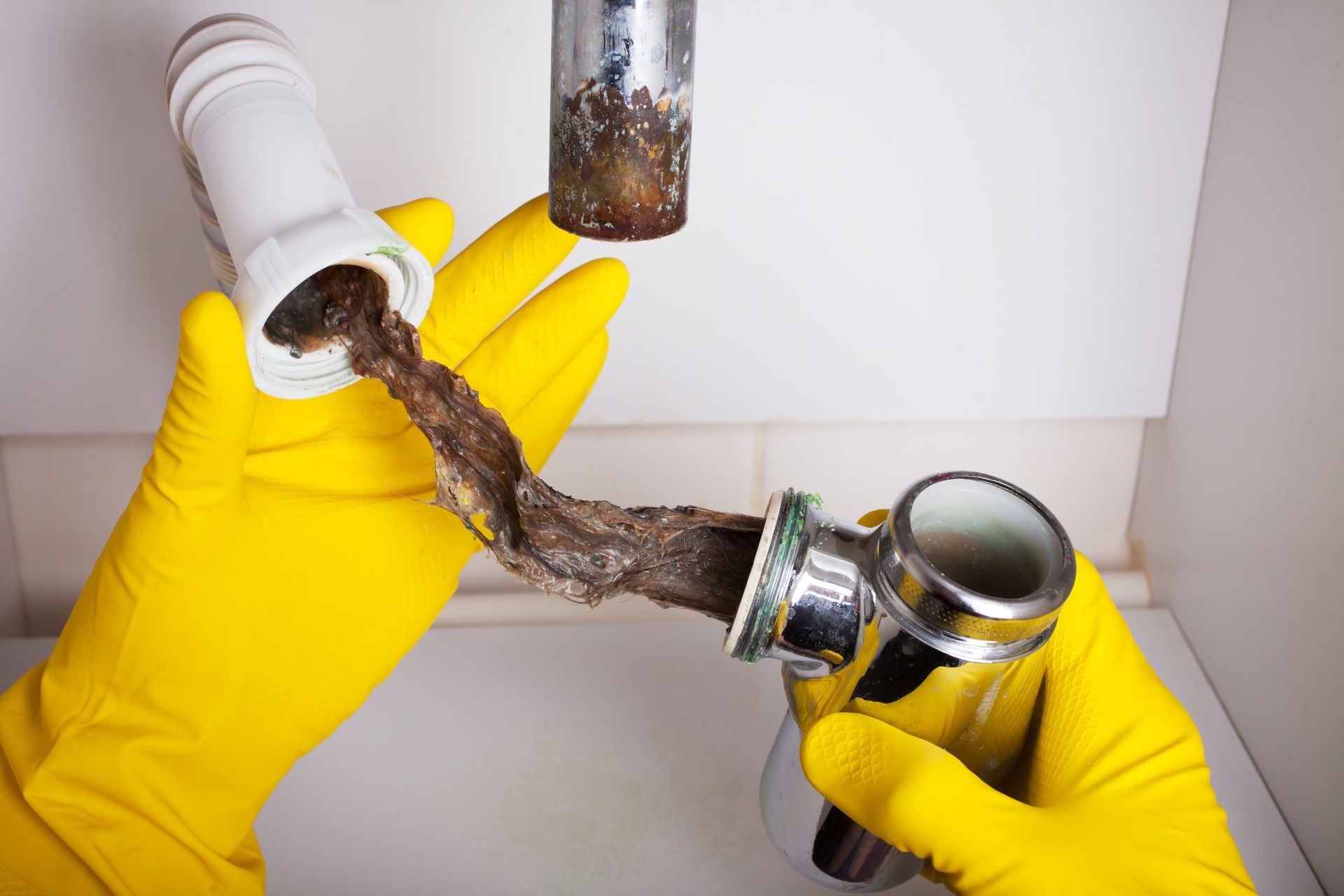 Yellow-gloved hands clear a clogged sink drain pipe under a white counter.