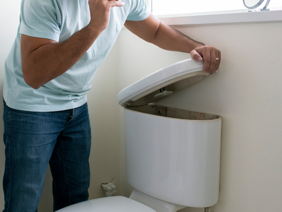 Man in jeans and a light blue shirt looking into an open toilet tank in a bathroom.