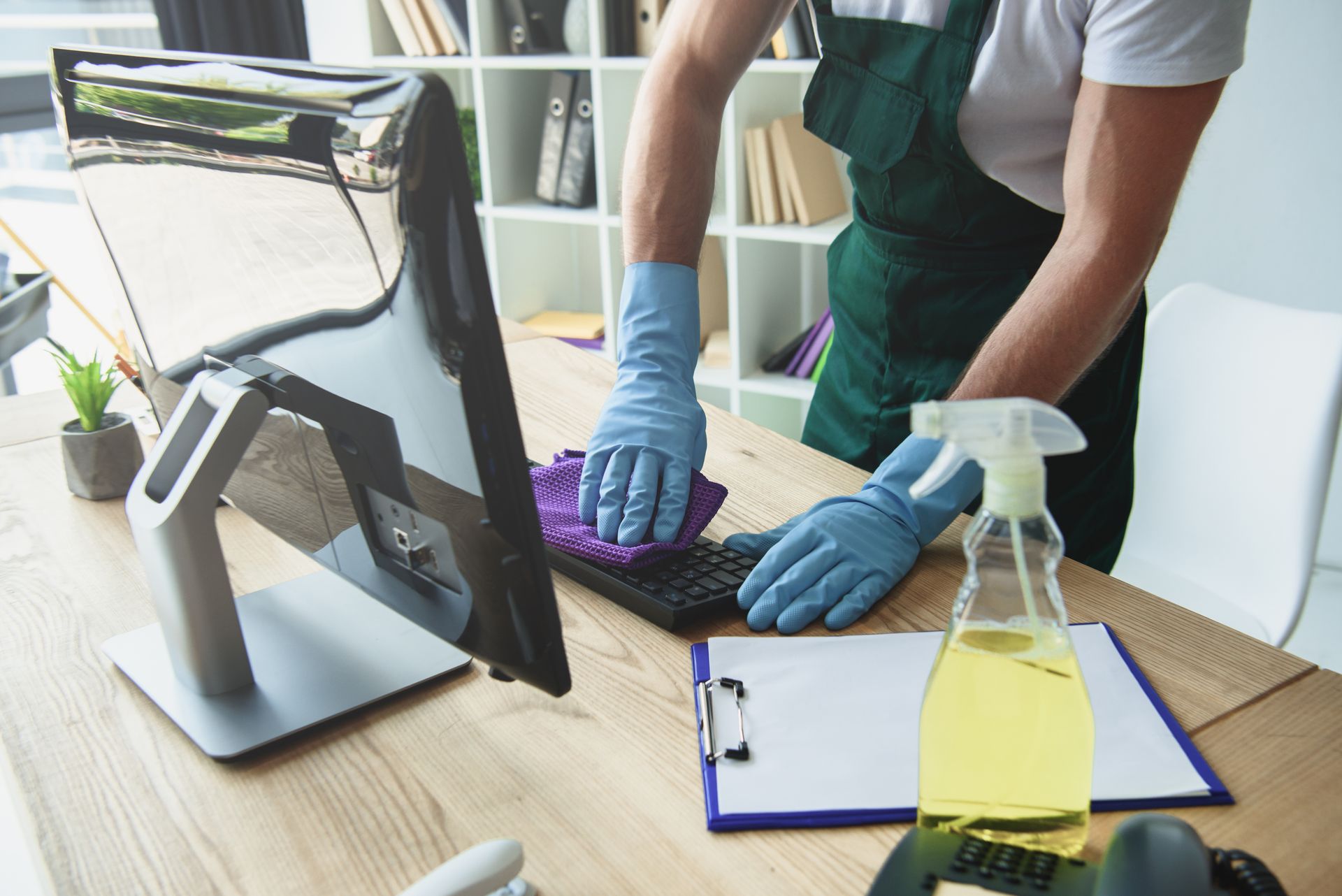 Person wearing gloves cleaning a computer keyboard with spray bottle on desk.