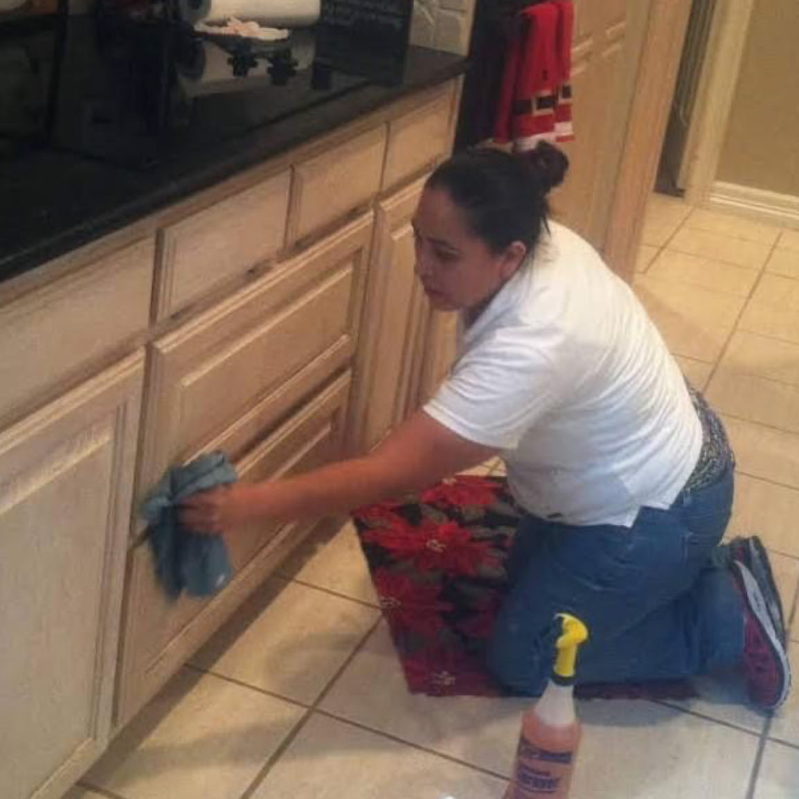 A person kneeling on a tiled floor and cleaning a light wood kitchen cabinet with a cloth and spray bottle.