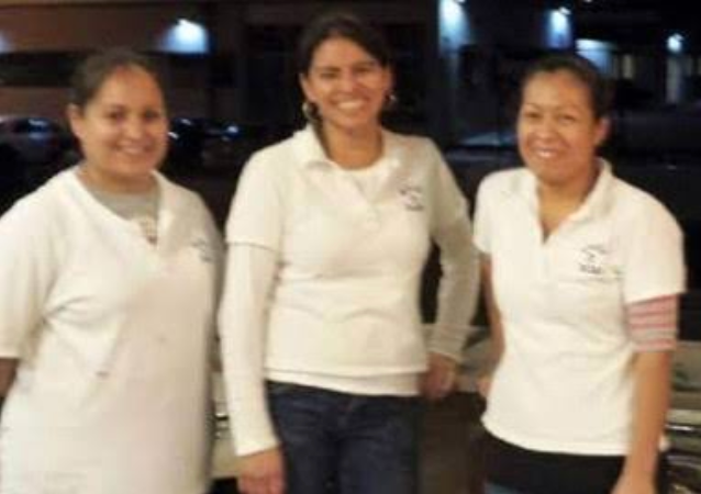 Three people in white shirts stand side-by-side smiling in a dimly lit outdoor setting.