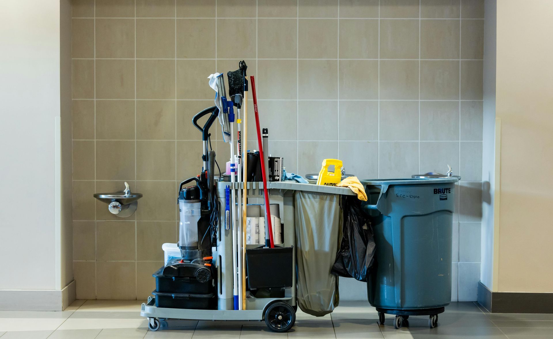 A janitorial cleaning cart loaded with supplies, a vacuum, and a large blue waste bin parked against a tiled wall.