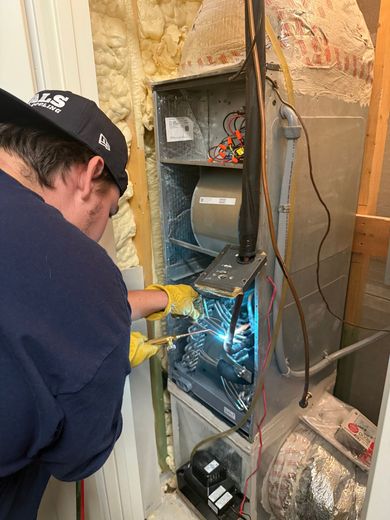 A person with a hat and gloves works on a furnace. The person is using a tool.