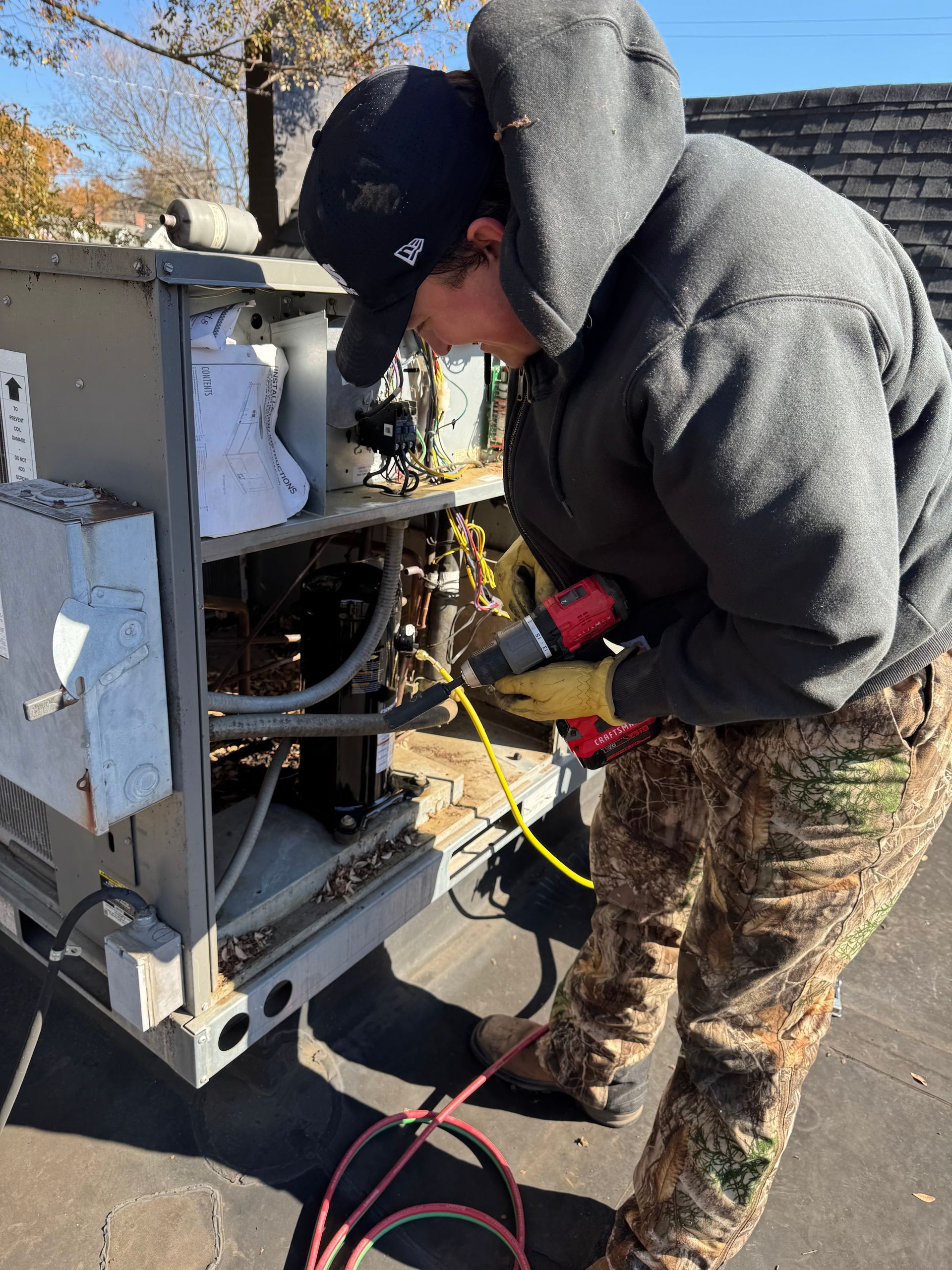 Person in camo pants and hoodie repairs an outdoor HVAC unit, using a drill.