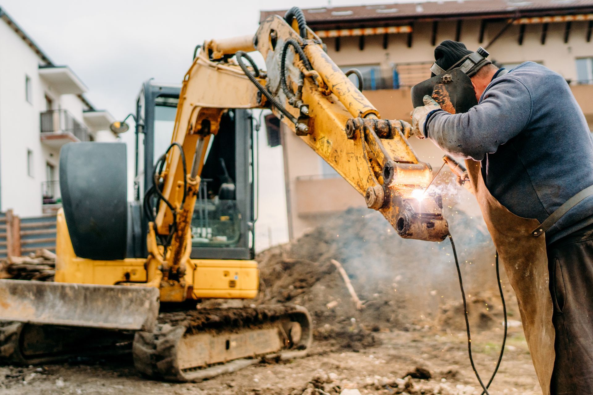 A person wearing protective welding gear repairs a yellow excavator arm on a construction site.