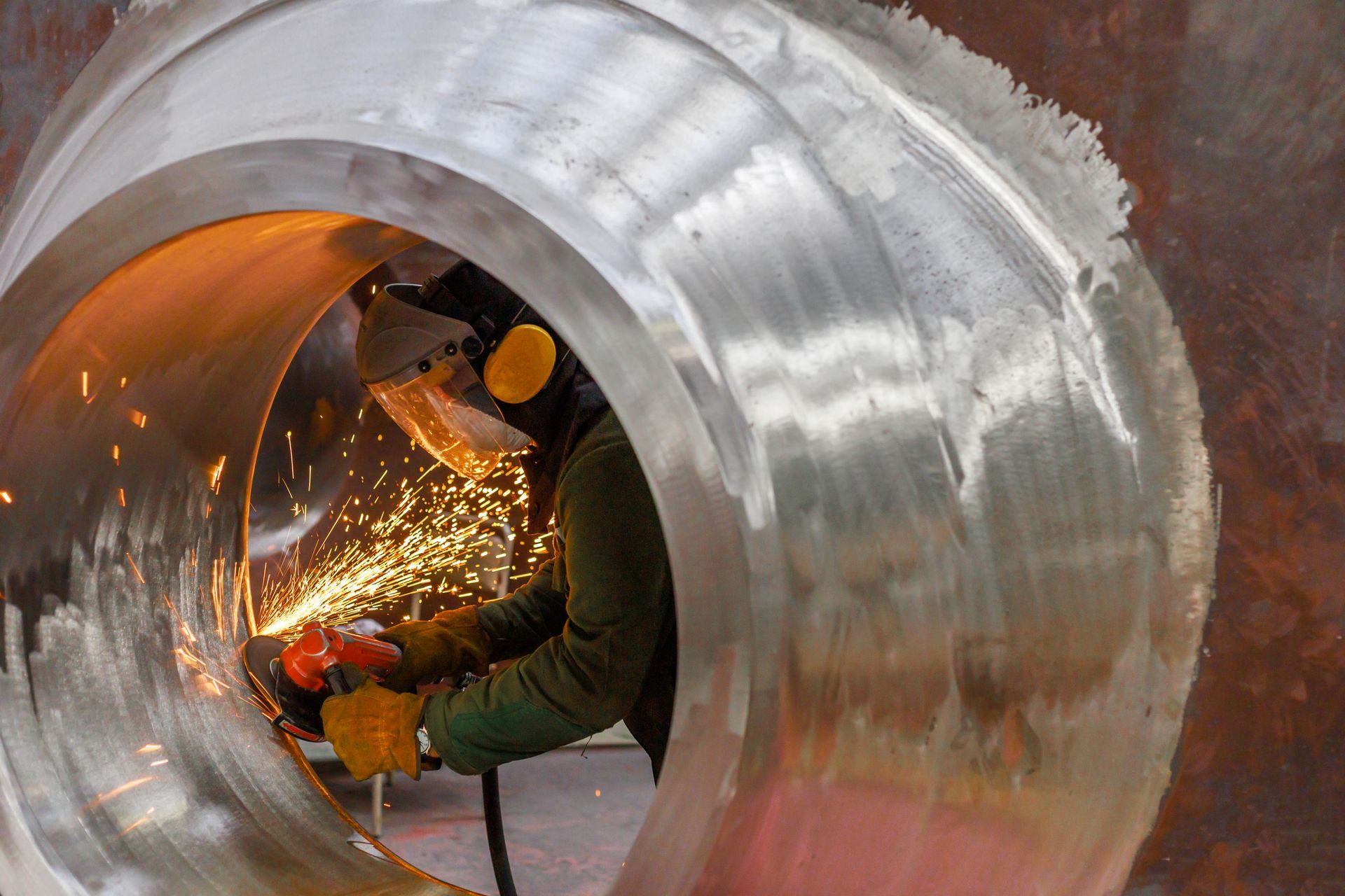 Worker using an angle grinder inside a large steel pipe, cutting metal as sparks fly.