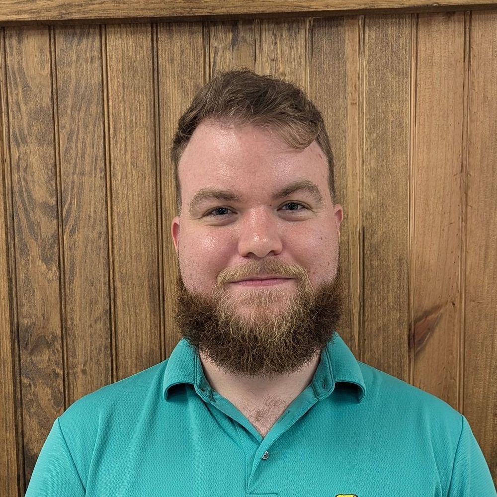 Man with a beard wearing a teal polo shirt, smiles in front of a wood-paneled wall.
