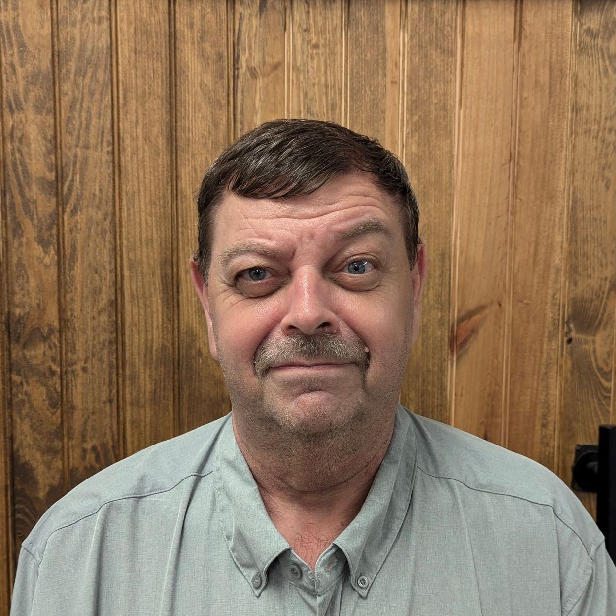 Man with short brown hair and mustache in a blue button-down shirt, standing in front of wood paneling.