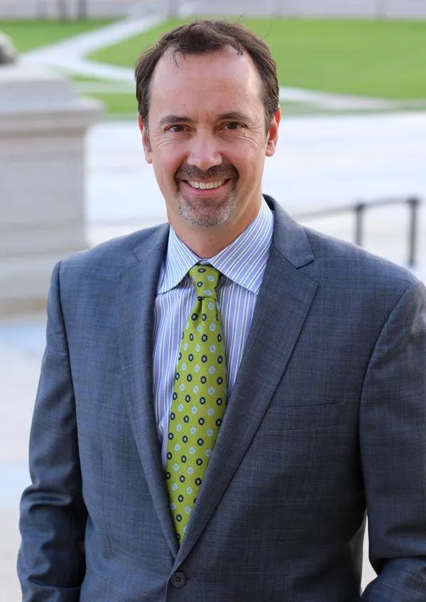 Smiling man in a gray suit and green tie standing outdoors near a walkway