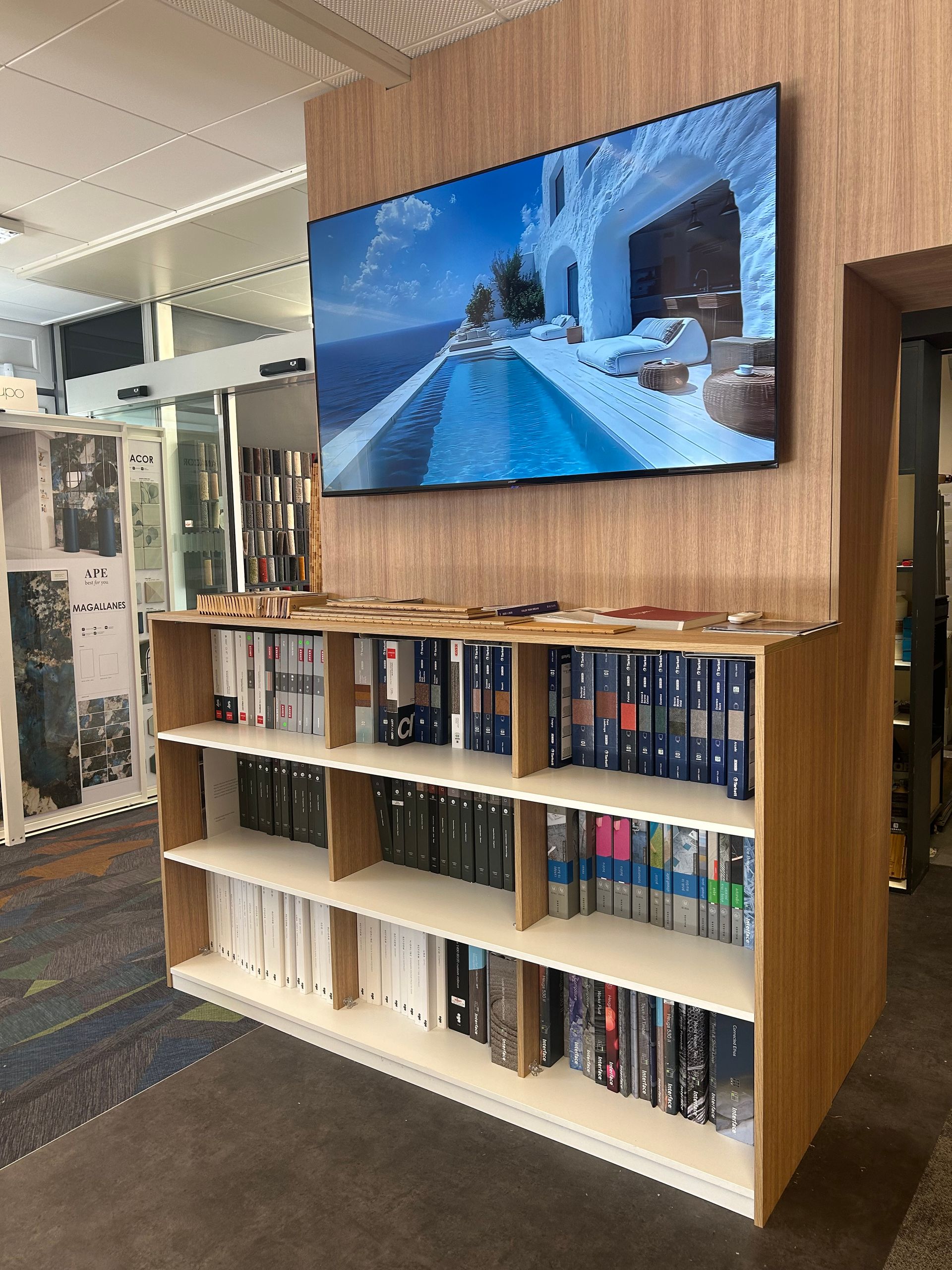 Une étagère en bois remplie de classeurs, placée sous un téléviseur mural diffusant des images d'une station balnéaire.