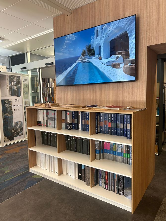 Une étagère en bois remplie de classeurs, placée sous un téléviseur mural diffusant des images d'une station balnéaire.
