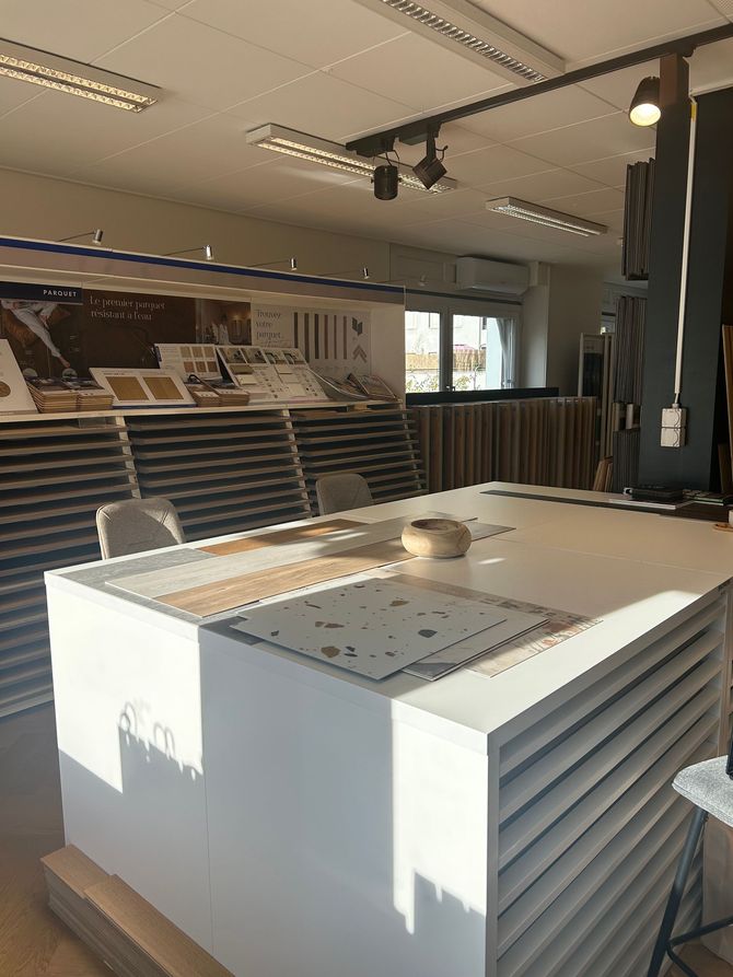 Une salle d'exposition avec une grande table centrale blanche présentant des échantillons de revêtements de sol devant des présentoirs de carrelage et de stratifié.