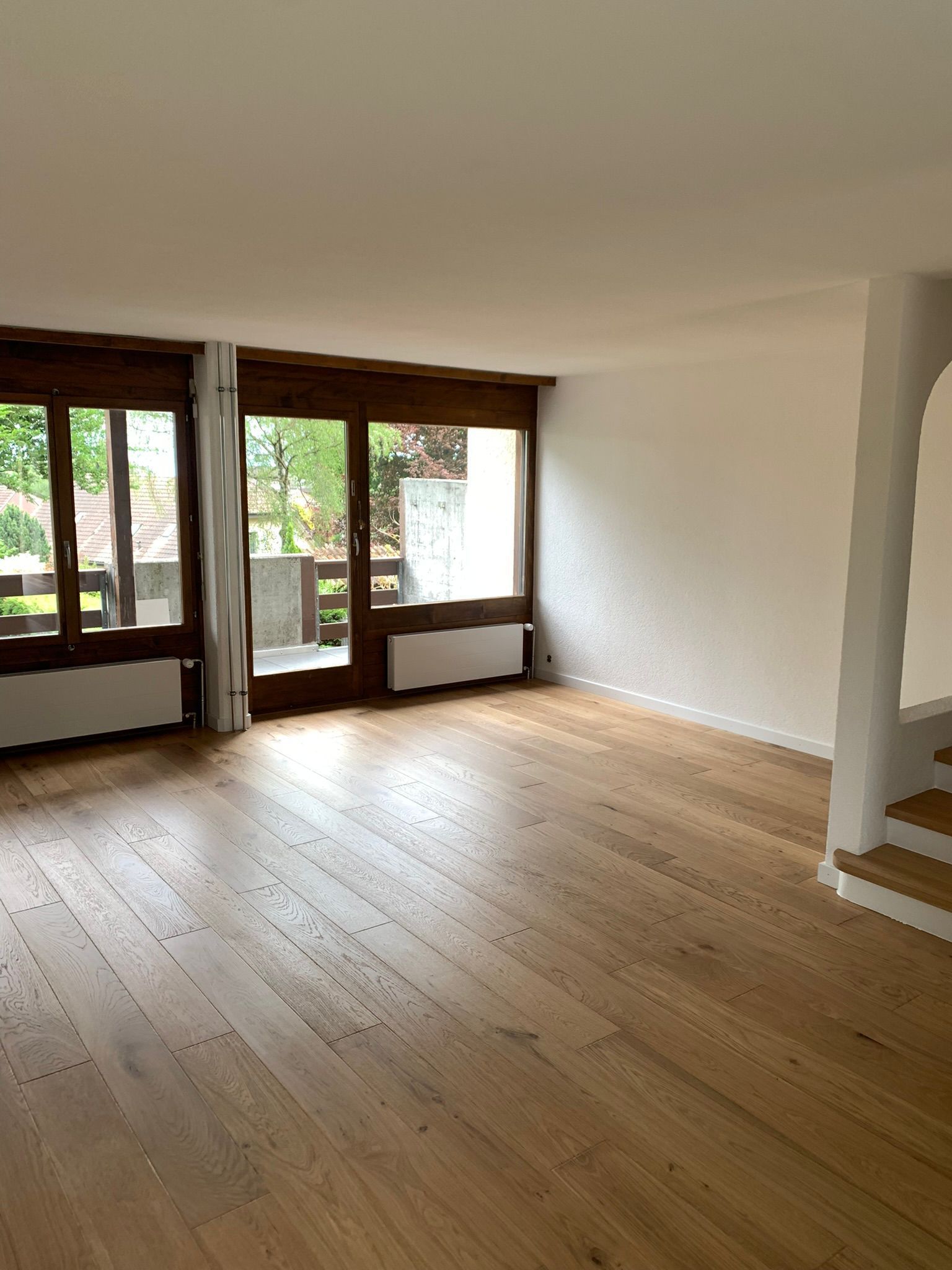 Salon vide avec parquet clair, murs blancs et grandes baies vitrées donnant sur un balcon.