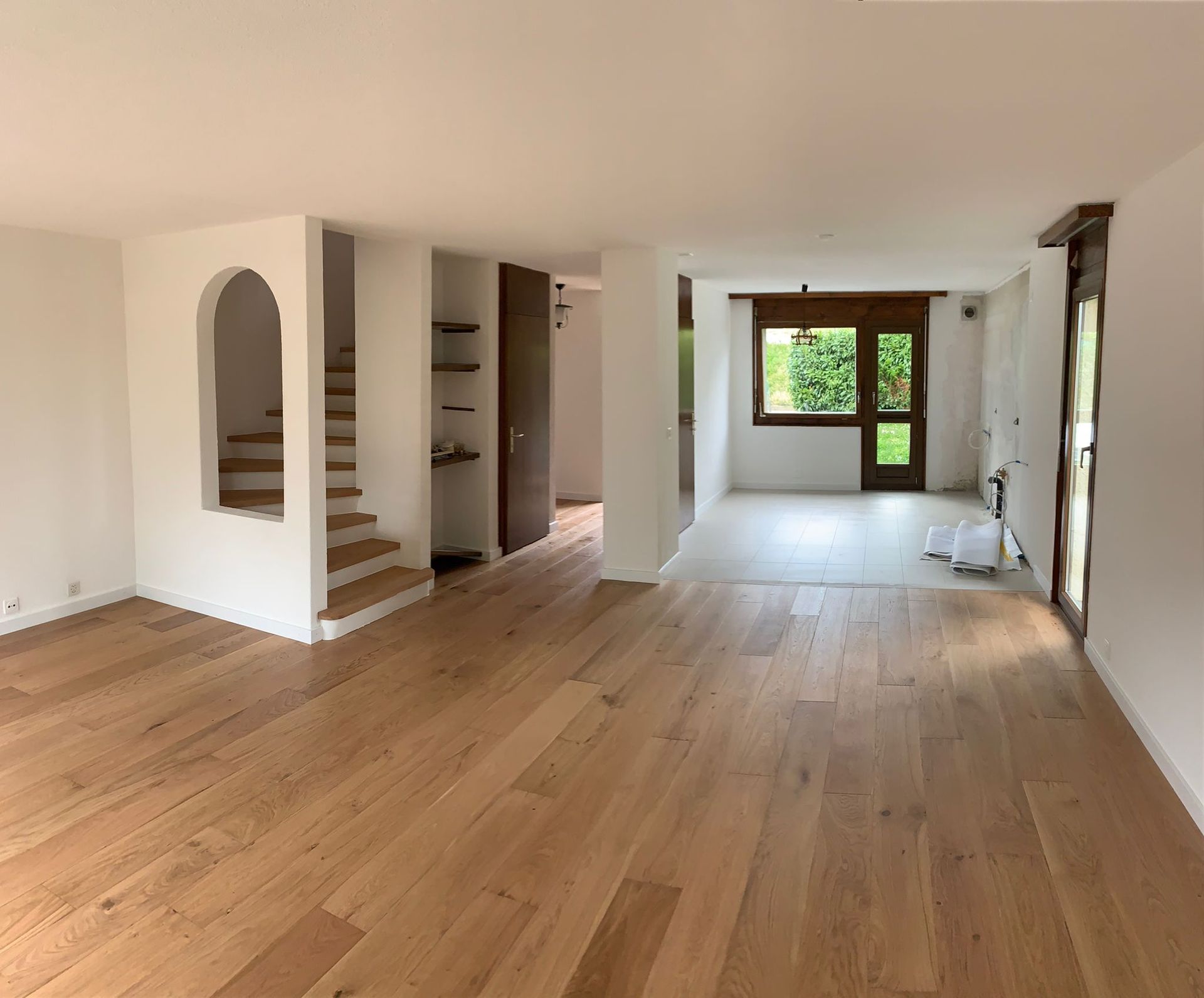 Une pièce vide et rénovée, avec un parquet clair, des murs blancs, un escalier voûté et une porte donnant sur un espace baigné de soleil.
